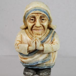 May include: A figurine of a smiling woman in a white and blue habit, with hands clasped in prayer. The figure has a serene expression and is wearing a head covering. The base of the figurine is dark gray.