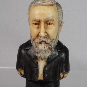 May include: A ceramic figurine of a man with a white beard and gray hair. He is wearing a black suit jacket with two buttons. The figurine has a detailed face with brown eyes and a neutral expression. The figure is approximately 5 inches tall.