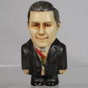 May include: A ceramic figurine of a man in a black suit, white shirt, and a red and green ribbon. The figurine has a smiling face with detailed facial features and dark hair. The figure is standing upright.