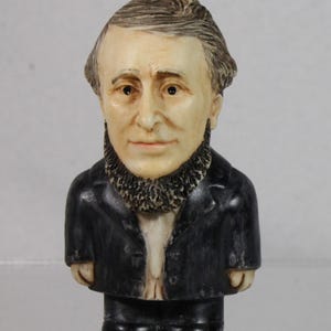May include: A small figurine of a man with a beard, wearing a black suit and a white shirt. The figure has a detailed face with brown hair and eyes. The figurine is standing upright.