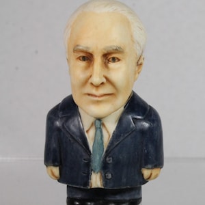 May include: A small figurine of a man with white hair, wearing a dark blue suit, a light blue tie, and a white collared shirt. The figurine has a detailed face and is standing upright.