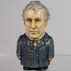 May include: A ceramic figurine of a man with gray hair and a stern expression. He wears a blue coat with gold buttons and a black tie. The figurine is a bust, with the head and shoulders visible.