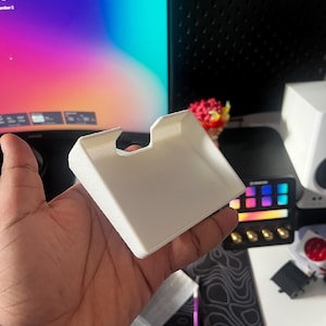 Może przedstawiać: Biały, prostokątny, drukowany w 3D przedmiot z zagłębionym obszarem i zakrzywioną górną krawędzią. Przedmiot jest trzymany w dłoni, z monitorem komputera i innymi akcesoriami technicznymi w tle.