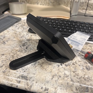 May include: A black electronic device is mounted on a black stand. The device is rectangular with a flat top and angled sides. The stand has a base and an arm that supports the device. The background includes a keyboard and a countertop.