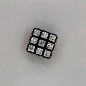 Può includere: Un cubo di Rubik's nero con scintillanti accenti di cristallo su ogni quadrato. I bordi sono rifiniti con gemme rosse, blu e verdi. Il quadrato centrale presenta un logo colorato.