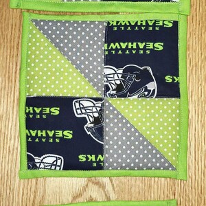 Pode incluir: Três bases de tecido quadradas com borda verde. Cada base apresenta um painel azul marinho com um capacete do Seattle Seahawks e o nome da equipe. O design inclui padrões de bolinhas verdes e cinza.