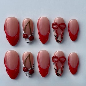 May include: Set of ten artificial nails with a light pink base and red tips. Some nails feature cherry designs, bow designs, and black and white stripes. The nails are oval-shaped.