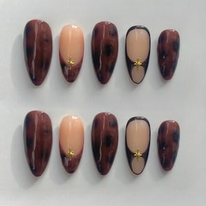 May include: A set of ten oval false nails. The nails feature a tortoiseshell design in brown and black, with some having a nude base and a gold star accent. The nails are arranged in two rows of five.