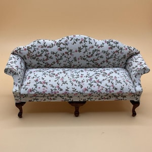 May include: A miniature sofa with a floral design. The sofa has a white base with pink and green floral details. It features a curved back and armrests, and is supported by four dark brown legs. Ideal for a doll's house or miniature display.
