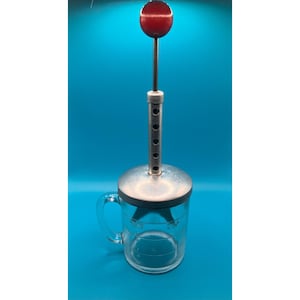 May include: A clear glass measuring cup with a metal lid and a vertical plunger mechanism. The plunger features a thin metal rod topped with a round red knob. The glass container has a side handle and printed measurement markings on the exterior.