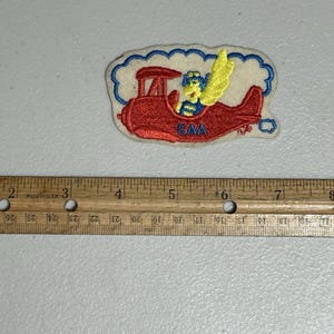 May include: A red aeroplane with a yellow and blue cartoon character flying in the clouds. The aeroplane has the letters "EAA" on the side.