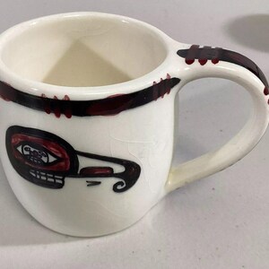 May include: White ceramic mug with a black and red design. The design features a stylized bird with a red eye and a black and red border around the top of the mug.