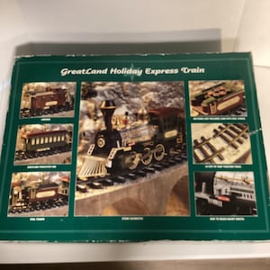 May include: A green box showing a GreatLand Holiday Express Train set. The box has images of a black and gold locomotive, passenger carriages, coal tender, and track. The train set is designed to run on a track.