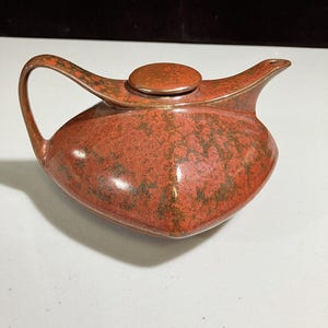 May include: A ceramic teapot with a lid and handle. The teapot is a reddish-orange color with dark green speckles. The handle curves up and around, and the spout is small and pointed.