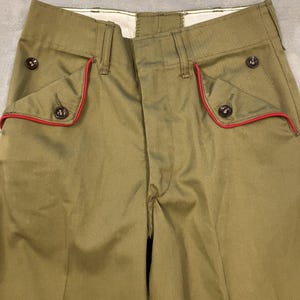 May include: Olive green pants with red trim around the pockets and dark buttons. The pants have a classic design with a button closure and belt loops. The fabric appears to be a durable material, suitable for everyday wear.