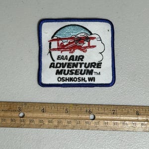 May include: A white embroidered patch featuring a red aeroplane flying over a blue and white cloud. The text "EAA AIR ADVENTURE MUSEUMTM OSHKOSH, WI" is embroidered in black.