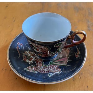 May include: A porcelain teacup and saucer set featuring a dark navy blue background with a raised, multi-colored dragon motif in red, green, and white. The cup has a metallic gold-toned handle, and both pieces are finished with a thin gold rim.