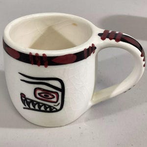 May include: White ceramic mug with a black and red design. The design features a stylized face with a red eye and black teeth.