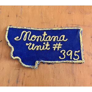 Peut inclure: Un écusson brodé bleu en forme de l'État du Montana avec des bordures cousues jaunes et un texte jaune indiquant Montana Unit # 395. L'écusson présente une surface en tissu texturé et est conçu pour être fixé sur des vêtements ou des accessoires.