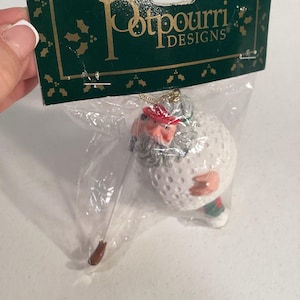 Peut inclure: Ornement de Noël sur le thème du golf, représentant un golfeur sortant d'une balle de golf. Le golfeur porte un chapeau rouge et a une barbe blanche. L'ornement est dans un sac en plastique transparent avec une étiquette verte "Potpourri Designs."