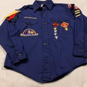 May include: A navy blue Cub Scouts BSA button-down shirt with long sleeves. The shirt features two chest pockets and several patches, including "Inland Empire Council" and various achievement badges. A colorful shoulder knot and patches adorn the sleeves.