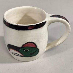 May include: White ceramic mug with a black and red band around the top and a green and red bird design on the front.