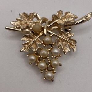 Könnte beinhalten: Goldfarbene Brosche in Form einer Traube. Die Brosche hat detaillierte Blätter und Stiele, mit Kunstperlen, die die Trauben darstellen. Ein klassisches, elegantes Accessoire.