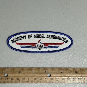 May include: An embroidered patch with the text "ACADEMY OF MODEL AERONAUTICS" in blue and white. The patch features a red and white design with the letters "AMA". The patch is approximately 6 inches long.