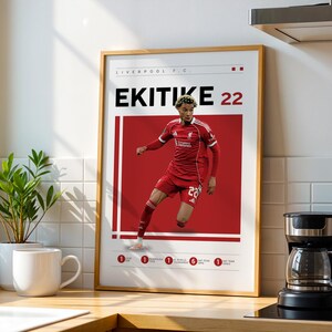 以下が含まれることがあります： 赤いユニフォームを着たサッカー選手が描かれた額入りのポスター。名前「EKITIKE 22」が大きく表示されています。赤と白のデザインで、リバプールFCのロゴも含まれています。