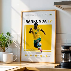 May include: A framed poster featuring a football player in a yellow and green jersey with the text AUSTRALIA NATIONAL SOCCER TEAM, IRANKUNDA, and the number 17. The design includes a yellow rectangular background and small circular icons at the bottom.