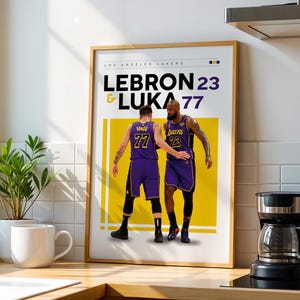 May include: A framed poster featuring two basketball players in purple and gold uniforms. The text "LOS ANGELES LAKERS" is at the top, with "LEBRON 23 & LUKA 77" below. The poster is displayed on a light-colored wall.