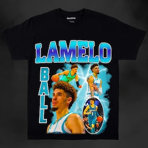 以下が含まれることがあります： バスケットボール選手のラメロ・ボールのグラフィックプリントが施された黒いTシャツ。グラフィックは青と緑のカラーリングで、稲妻のデザインが施されています。"LAMELO BALL"という文字が大きく太字で印刷されています。