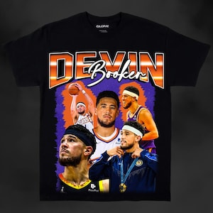 May include: Black t-shirt with a retro-style graphic featuring Devin Booker, a basketball player for the Phoenix Suns. The graphic includes Booker's name in large, colorful letters and images of him playing basketball. The shirt also features a PayPal logo.