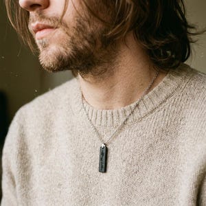 Peut inclure: Un collier en chaîne argentée avec un pendentif rectangulaire noir. Le pendentif porte un texte gravé et est suspendu à une chaîne délicate. La personne porte un pull beige.