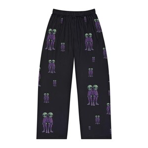 Op de afbeelding: Zwarte pyjamabroek met een patroon van paarse aliens. De aliens zijn in paren afgebeeld, gekleed in paarse outfits met groene hoofden en grote zwarte ogen. De broek heeft een wijde pijp en een elastische tailleband.