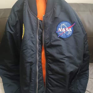 May include: A navy blue bomber jacket featuring a circular NASA patch on the left chest. The jacket has a full-length silver metal zip, a ribbed collar, and a bright orange quilted lining visible through the open front. A yellow patch is on the sleeve.