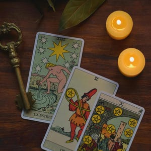 May include: A collection of tarot cards with illustrations, including a card with a nude figure and a star, and a card with a figure holding coins. A golden key and two lit candles are also present.