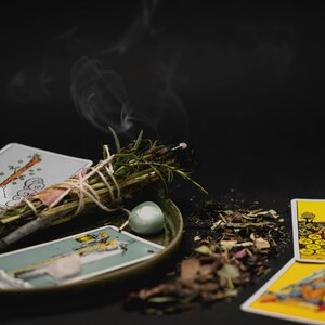 May include: A still life arrangement featuring tarot cards, crystals, and a burning sage bundle on a dark surface. The cards have colorful illustrations, and smoke rises from the sage. The scene suggests a spiritual or mystical theme.