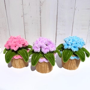 Può includere: Tre decorazioni floreali per cupcake fatte a mano. Ogni cupcake presenta un fiore di colore diverso: rosa, lavanda e blu. I fiori poggiano su foglie verdi e sono inseriti in un pirottino marrone con bordo smerlato.