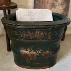 May include: A dark green, antique planter with gold floral designs. The planter has a wide, round shape and is filled with a piece of white fabric. In the background, there is a wooden cutting board and a small wooden stool with a brown ceramic vase.