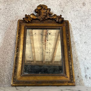 May include: A rectangular wall mirror with a weathered gold-toned frame featuring a decorative carved crest at the top. The glass surface shows signs of age with visible dark spots and a reflective quality that captures the surrounding space.
