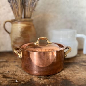 May include: A copper pot with a lid and gold-colored handles sits on a wooden surface. The pot reflects the light, showcasing its polished surface. A ceramic vase with dried lavender and a white mug are in the background.