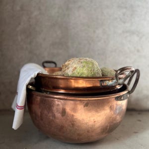 Peut inclure: Trois bols en cuivre antiques de différentes tailles, empilés les uns sur les autres. L'un des bols contient un légume vert clair et un tissu blanc à rayures rouges. Les bols ont des poignées et un aspect vieilli et poli.