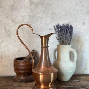 Könnte beinhalten: Eine Kupferkanne mit gebogenem Griff und ausgestelltem Ausguss. Die Kanne wird mit einem Holzstößel und -mörser sowie einer weißen Vase mit getrocknetem Lavendel präsentiert. Die Objekte stehen auf einer Holzoberfläche.