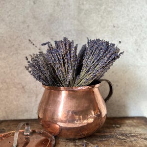 Könnte beinhalten: Ein Kupfertopf mit Griff und Deckel, gefüllt mit getrocknetem Lavendel. Der Topf hat eine gehämmerten Textur und steht auf einer Holzoberfläche. Der Lavendel hat violette Blüten und grüne Stiele.
