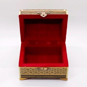 May include: An open wooden jewelry box with a red velvet interior. The box is rectangular with decorative gold and black floral patterns around the edges. A small metal clasp secures the lid.