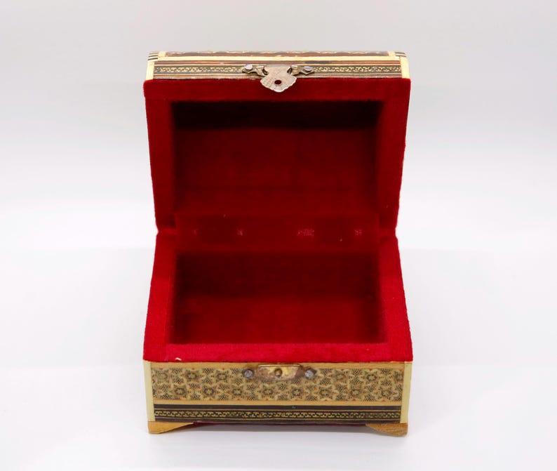 May include: An open wooden jewelry box with a red velvet interior. The box is rectangular with decorative gold and black floral patterns around the edges. A small metal clasp secures the lid.
