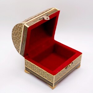 May include: An open, ornate wooden chest with a red velvet interior. The exterior features intricate mosaic patterns in shades of beige, brown, and gold. A small metal clasp secures the lid. The chest is elevated on small, decorative feet.
