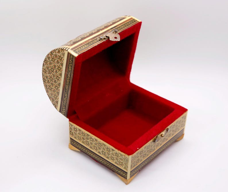 May include: An open, ornate wooden chest with a red velvet interior. The exterior features intricate mosaic patterns in shades of beige, brown, and gold. A small metal clasp secures the lid. The chest is elevated on small, decorative feet.