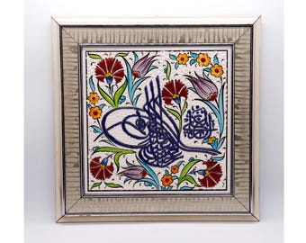 May include: A framed ceramic tile featuring a floral design with red carnations, purple tulips, and orange flowers. The tile has a central blue calligraphy design. The frame is silver.